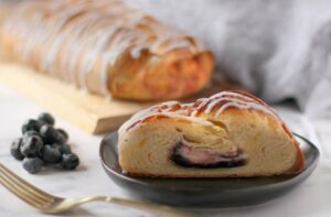 blueberry butter braid pastry slice with icing on plate with full pastry in back on cutting board with icing. Blueberries for garnish