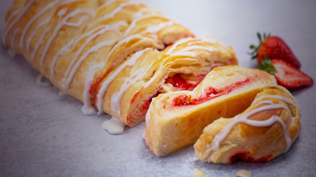Strawberry Cream Cheese Butter braid Pastry partially cut with icing and strawberries for garnish